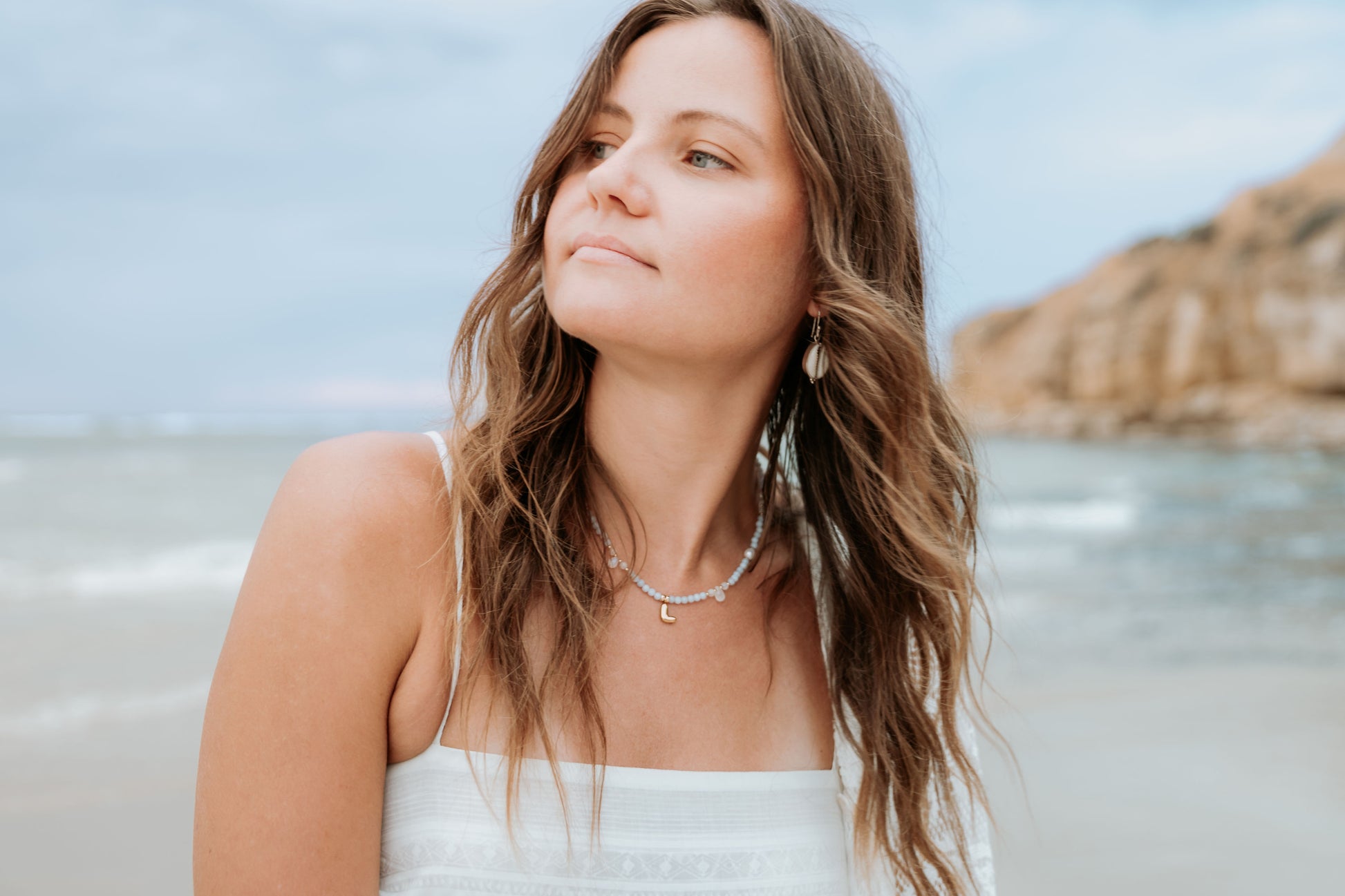 Woman with long hair standing on a beach with water and rocks in the background